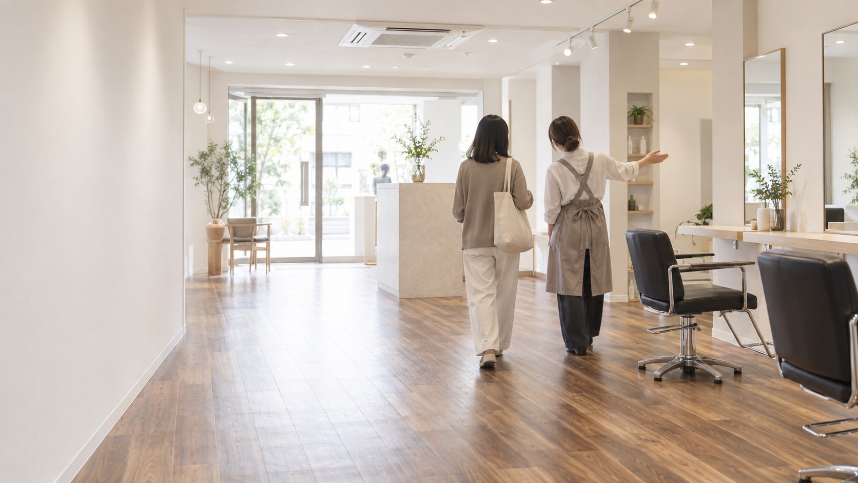 私服の人とエプロン姿のスタイリストが、明るい美容室の中を入口のほうへ歩きながら店内を見ている後ろ姿