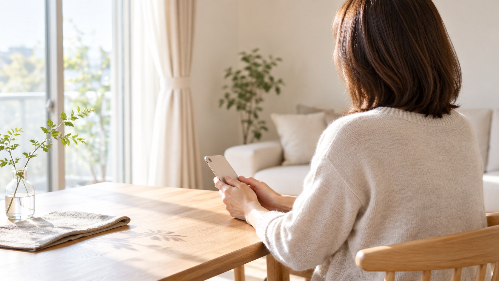 自宅の窓辺のテーブルで、女性がスマートフォンを両手に持っている後ろ姿